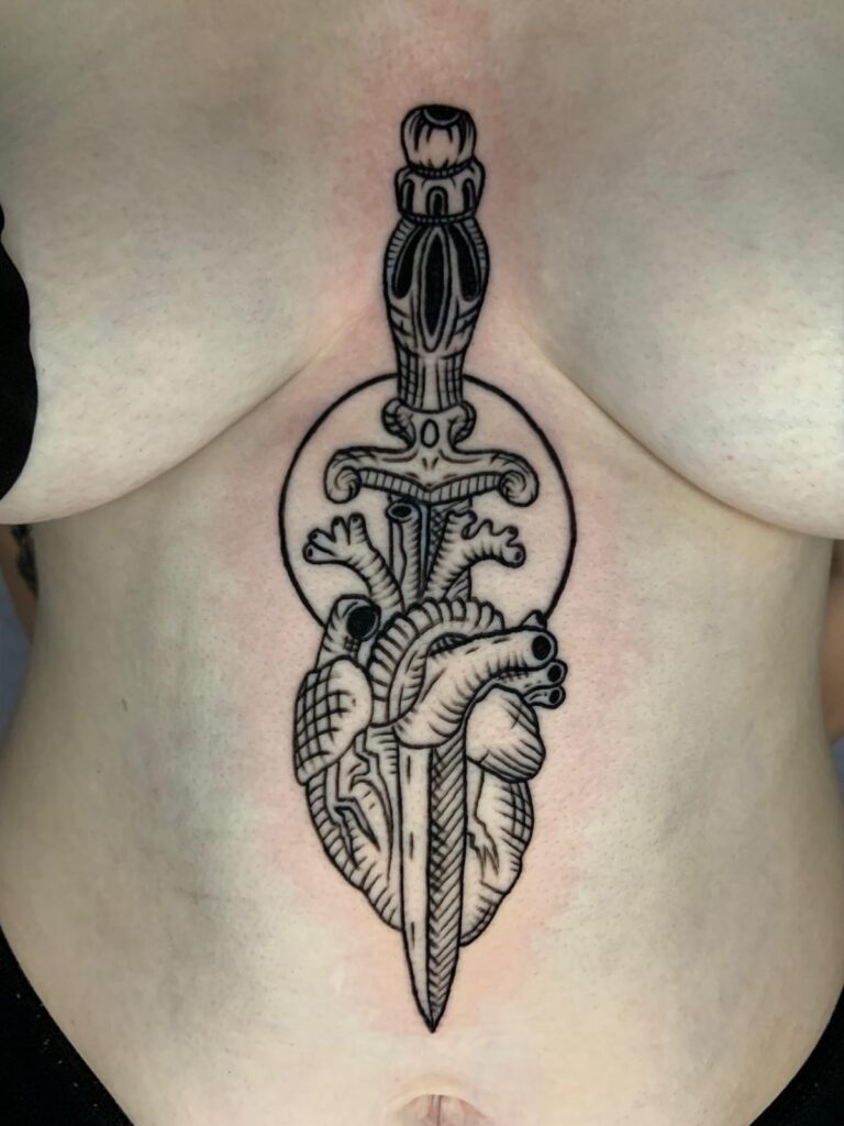 A symmetrical black-ink tattoo on a torso, positioned between the chest and navel. The design features a detailed, decorative dagger piercing vertically through an anatomical heart, set against a simple circular backdrop.