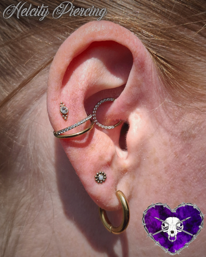 A detailed close-up of an ear showcasing a variety of gold and opal-accented jewelry by Milanda of Helcity Piercing. It features a crystal-lined daith hoop, an opal flower lobe stud, and a unique double-layered gold helix piercing.