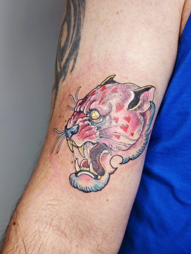 A vibrant, illustrative tattoo of a snarling pink big cat on an arm. The design features bright pink fur decorated with small red heart patterns, yellow eyes, and stylized blue shading along the mane.