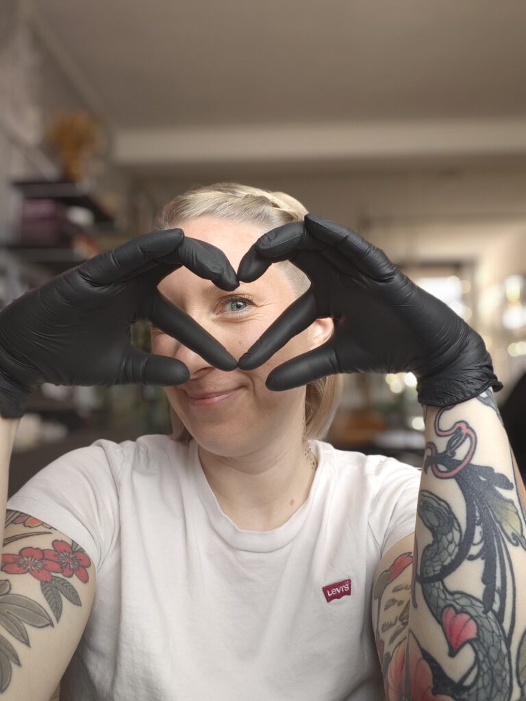 A portrait of tattoo artist Reinhilde from Electric Therapy. She is smiling warmly while wearing black tattooing gloves and using her hands to frame her eye in the shape of a heart. Her arms are adorned with vibrant, colorful illustrative tattoos.