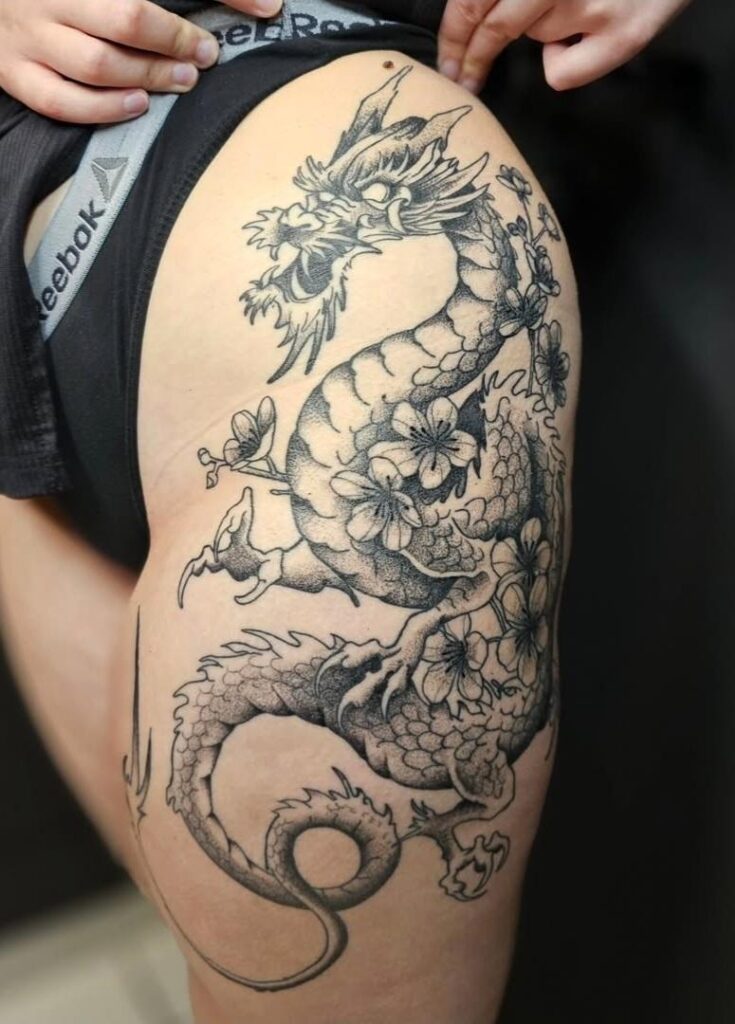 A large and intricate black and grey tattoo on a hip and thigh, created by Bryan from Mutant Ink. It depicts a winding traditional dragon woven between delicate cherry blossom branches with fine dot-work shading.