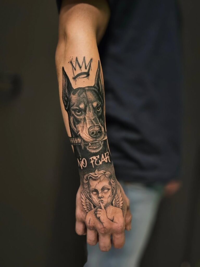 A multi-part black and grey tattoo on a forearm and hand by Bryan of Mutant Ink. The forearm shows a Doberman Pinscher wearing a crown above the text "NO FEAR," while the hand features a detailed cherub making a silencing gesture.