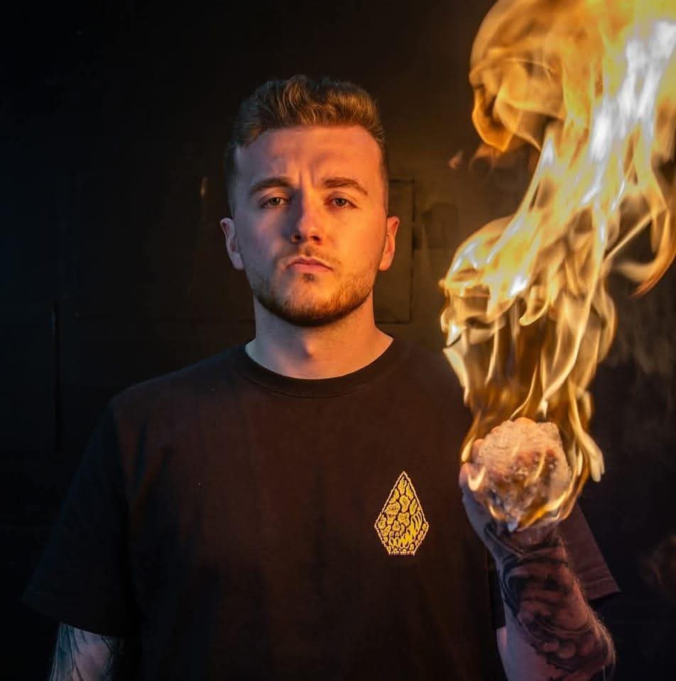 A dramatic portrait of tattoo artist Bryan from Mutant Ink. He is wearing a black t-shirt and holding a ball of bright, glowing flames in his tattooed hand against a dark, moody background.