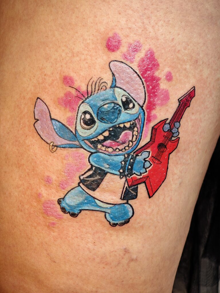 A bright and energetic tattoo of the character Stitch from Disney's Lilo & Stitch. He is depicted wearing a black leather vest and a spiked wristband while playing a red electric guitar against a pink watercolor-style splash background.