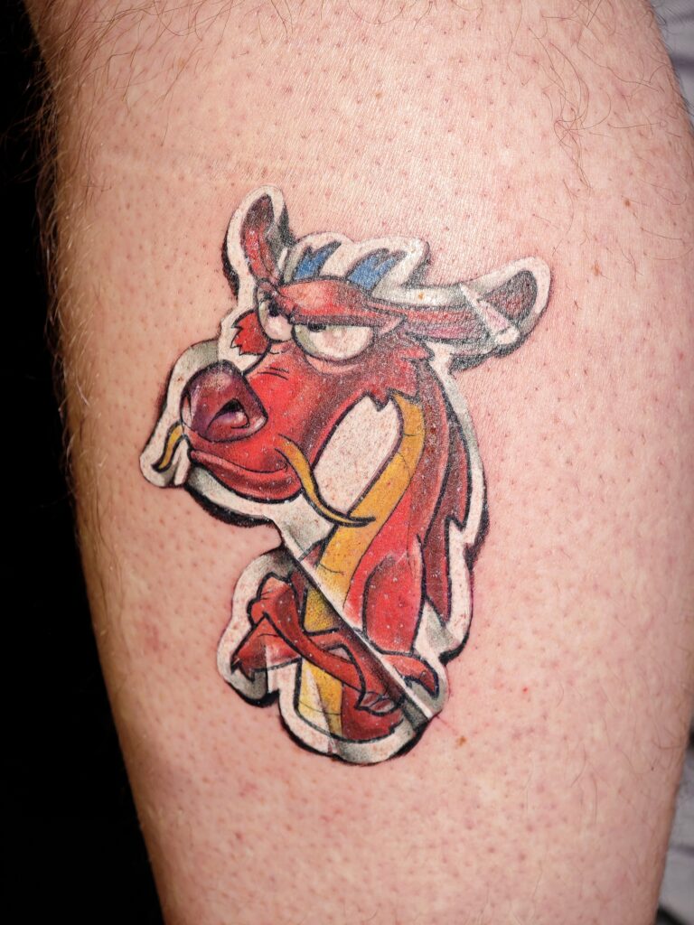 A colorful, sticker-style tattoo of the red dragon Mushu from Disney’s Mulan. The character is shown with a grumpy expression, rendered with bold black outlines and bright red and yellow saturation.
