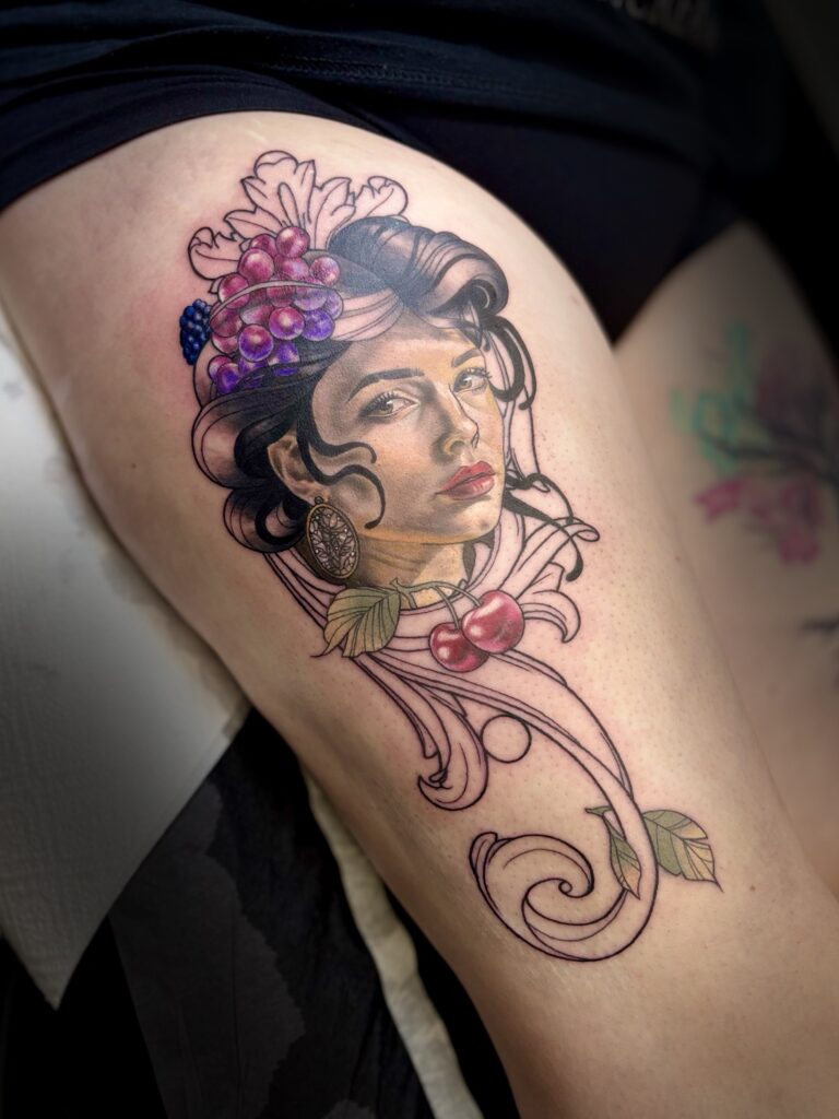 A vibrant, illustrative color tattoo on a thigh by Veerle from Skin Culture. It depicts a woman's face framed by clusters of purple grapes and red cherries, set against an elegant, flowing ornamental scrollwork background.