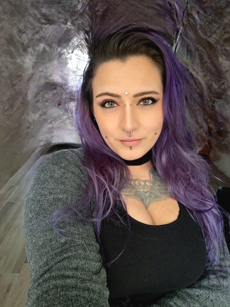 A portrait of piercing artist Milanda from Helcity Piercing. She has long, wavy purple hair and showcases a symmetrical array of facial piercings, including bridge, forehead, septum, cheek, and vertical labret piercings.