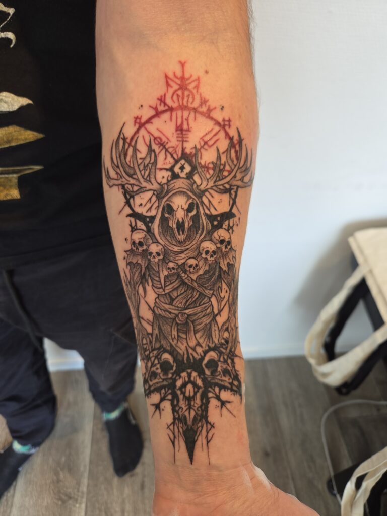 A striking black-ink tattoo on a forearm by Freyja of Deadly Muse. The design features a cloaked, skeletal figure with large antlers and a crown of small skulls, set against a dark, circular rune-like background with red accents.