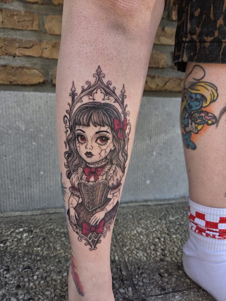 A detailed illustrative tattoo on a leg by Freyja from Deadly Muse depicting a gothic doll with cracked porcelain-like skin and large red eyes. The doll is adorned with red bows and encased in an intricate, dark gothic ornamental frame.