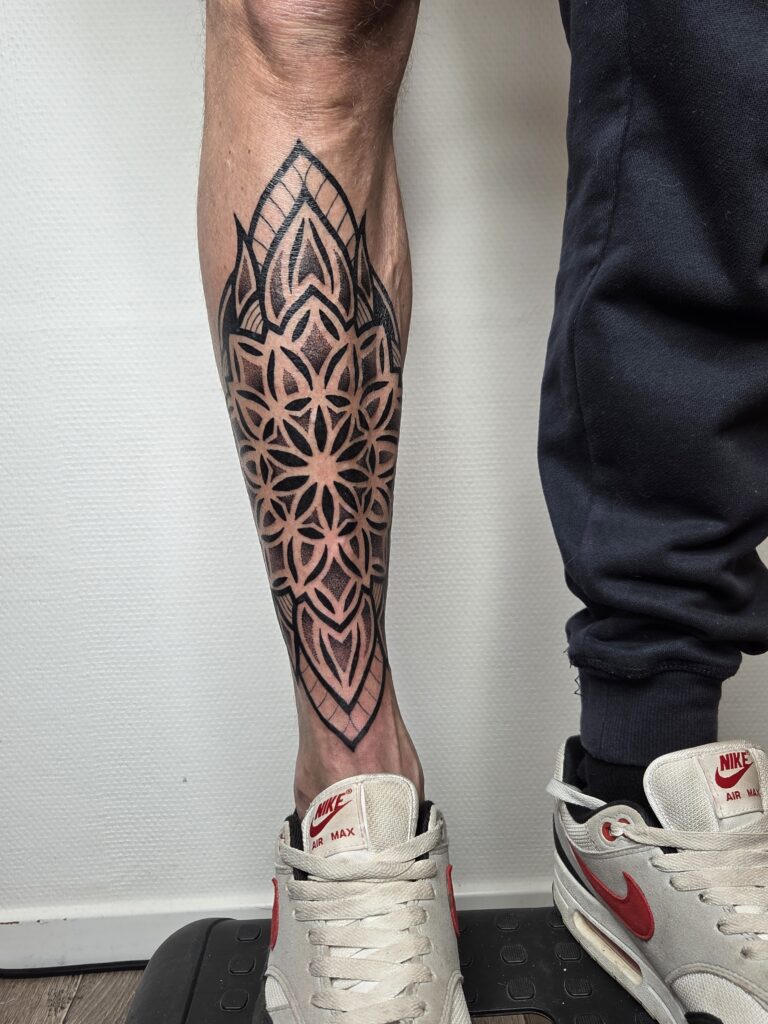 A bold, symmetrical geometric tattoo on a lower leg created by Freyja of Deadly Muse. The piece features a complex black-ink pattern inspired by the "Flower of Life," with sharp points and soft dotwork shading that follows the natural curve of the calf.