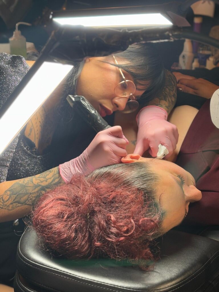 A close-up action shot of tattoo artist Celia from Poppy Lia focused on her craft. She is wearing glasses and pink gloves while carefully tattooing a delicate ornamental design behind a client's ear.