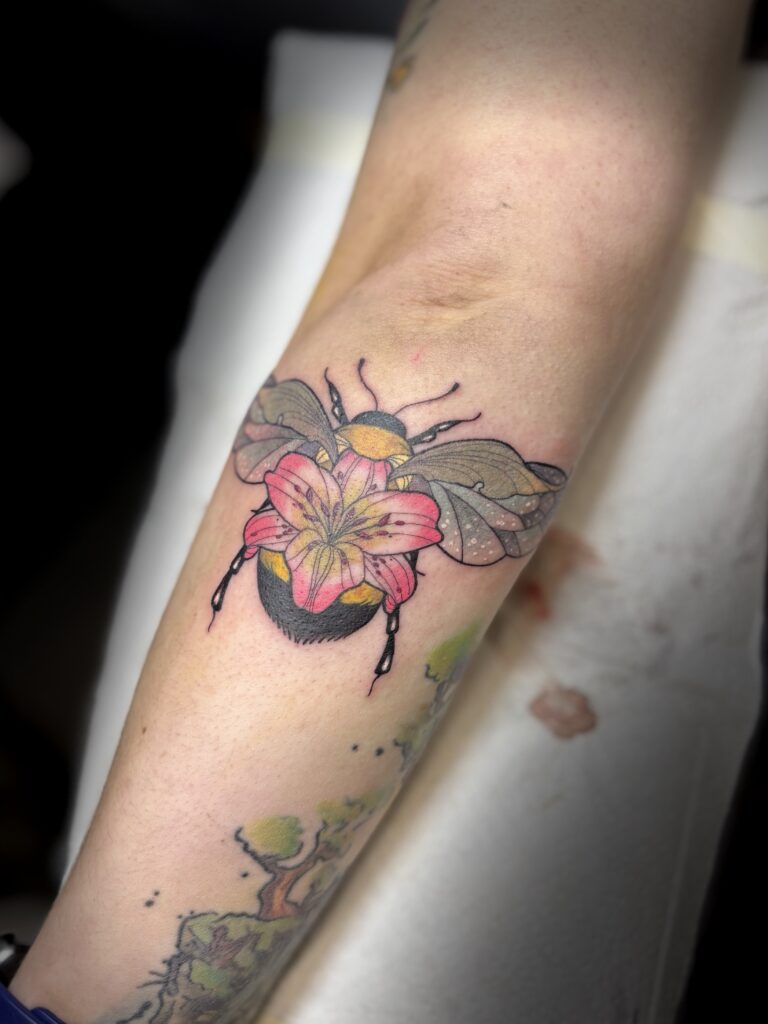 A detailed, nature-inspired tattoo on a forearm by Veerle of Skin Culture. It features a large bumblebee with a beautifully rendered pink lily flower growing from its back, showcasing her signature use of rich colors and soft shading.