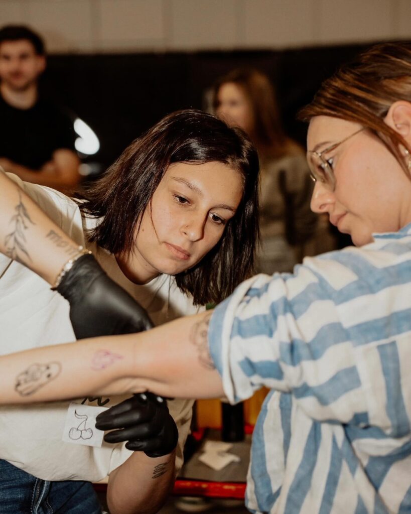 A candid shot of tattoo artist Laura from Inked By Laura focused on her work. She has dark hair, wears glasses, and is wearing black tattooing gloves while carefully applying a design to a client's arm.