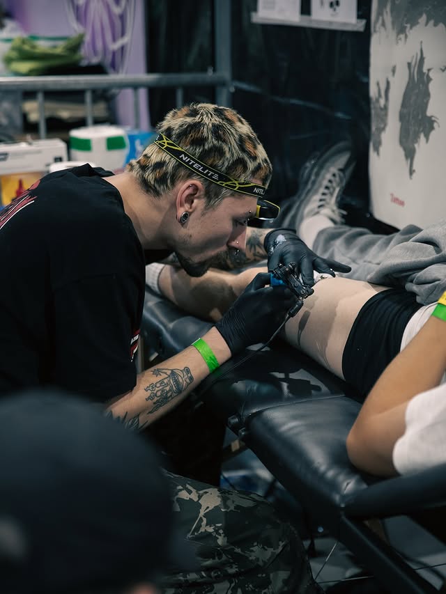 A candid shot of tattoo artist Ezra from TattooGast focused on his work. He has short hair with a distinctive leopard-print pattern and is wearing a headlamp while tattooing a client's leg in a studio setting.