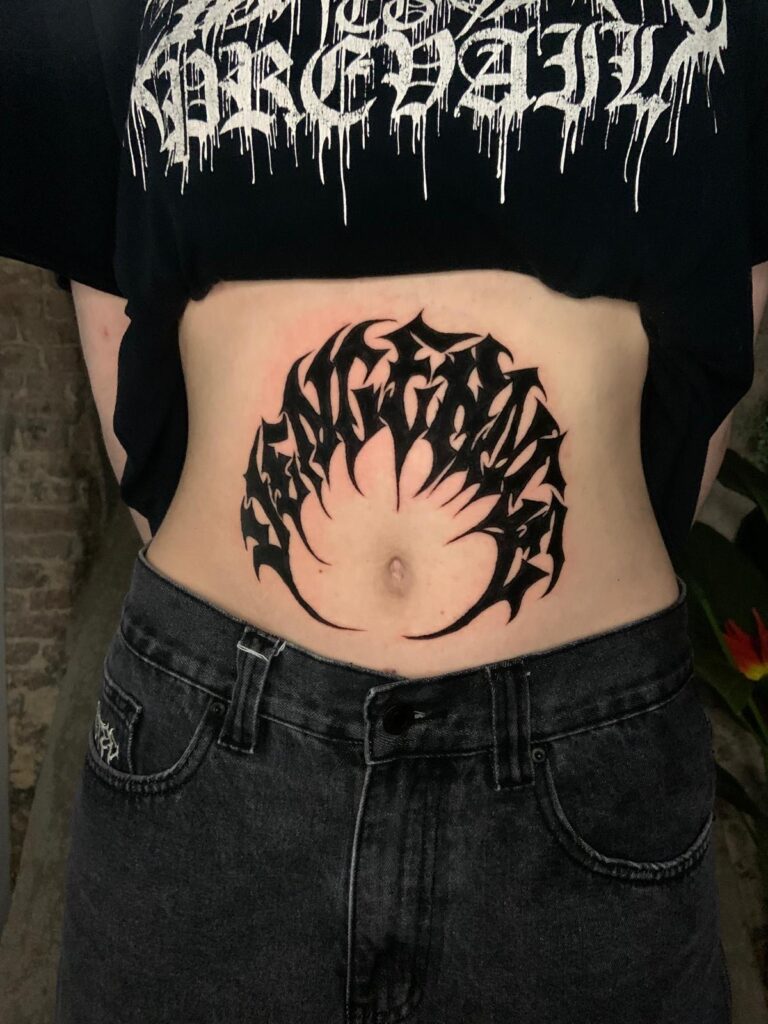A sharp, abstract black tattoo in a "cyber-sigilism" style, arched in a semi-circle above the navel. The person is wearing a black graphic t-shirt and dark denim jeans.