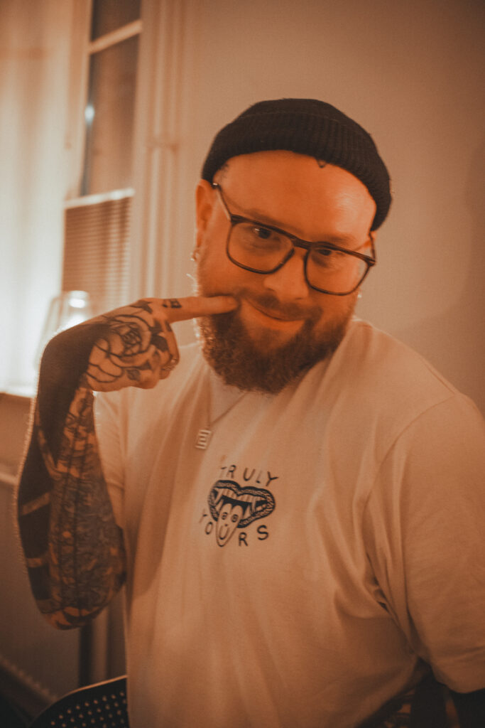 A portrait of tattoo artist Andy from Deux Tattoo. He has a full red beard and is wearing glasses, a black beanie, and a white t-shirt. He is smiling and pointing a tattooed finger toward his cheek.