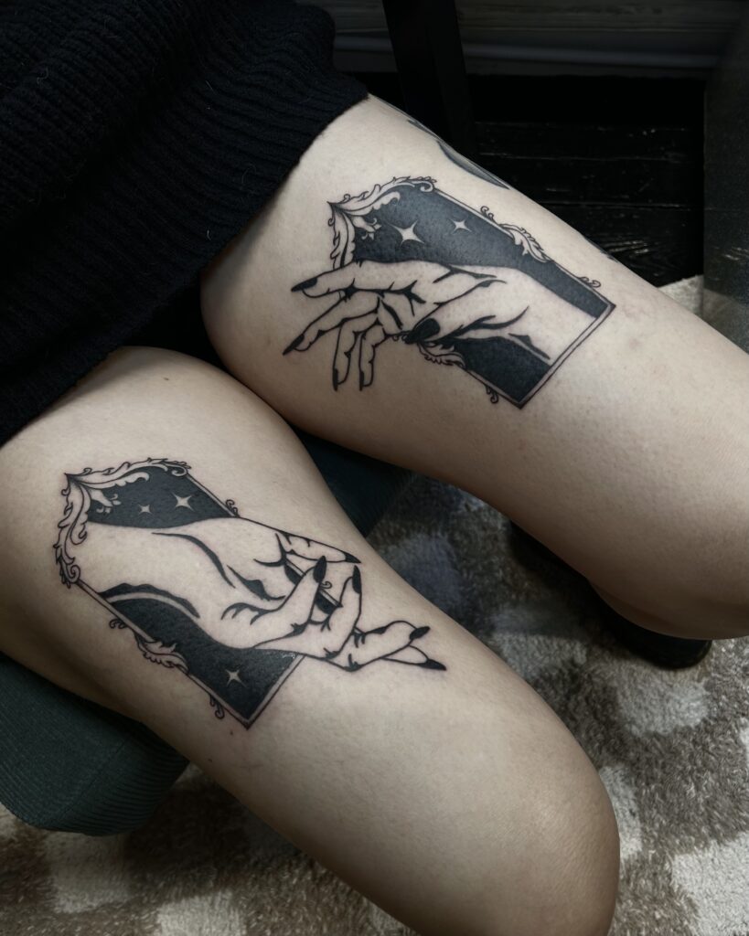 Symmetrical black-ink tattoos on a client's upper thighs by Helene of Donkerwerk. Each piece depicts a hand with long, dark nails emerging from a rectangular frame filled with a dark night sky and white stars.