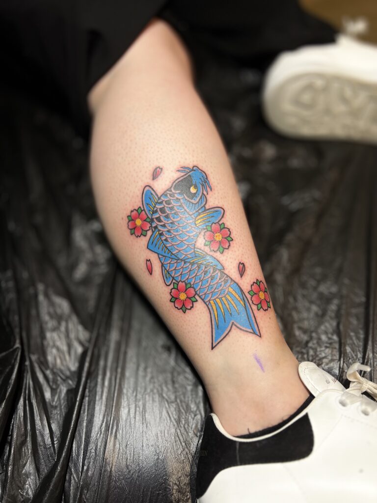 A vibrant, Japanese-style tattoo of a blue koi fish on a calf, swimming upward and surrounded by bright red cherry blossoms, created by Yento of Yentonic Tattoo.