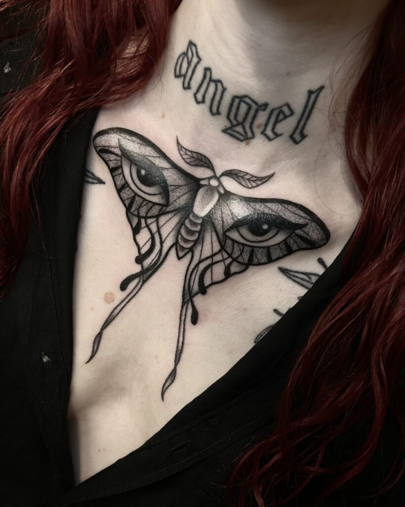 A bold, solid black-ink tattoo on a client's chest by Helene of Donkerwerk. The design features a large moth with detailed eyes on its wings, positioned directly below an existing "angel" tattoo on the throat.