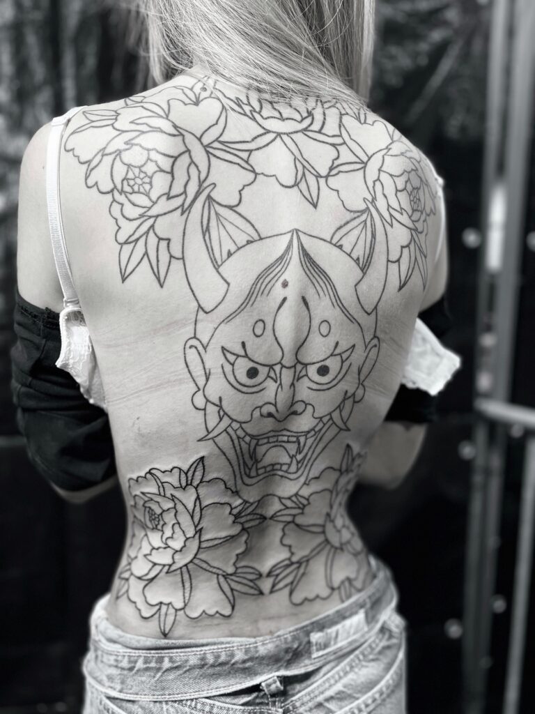 A large-scale back tattoo featuring an intricate Hannya mask centered between large blooming peonies. The detailed black-ink linework design is by artist Yento of Yentonic Tattoo.