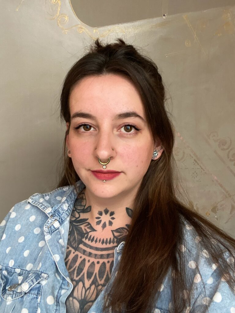 A portrait of tattoo artist Lavinja from LesDix Noirs, highlighting her intricate geometric black-ink neck and chest tattoos and her gold septum piercing.