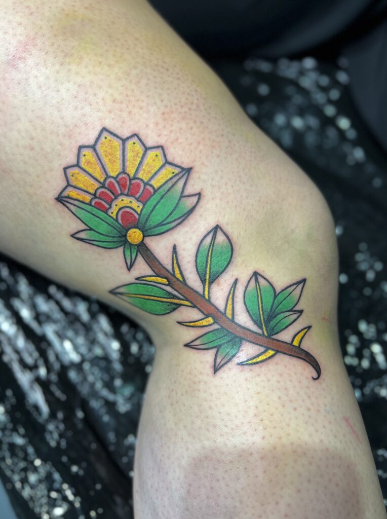 A colorful, traditional-style tattoo of a stylized yellow and red flower with a thorny green stem, showcasing the clean linework and bold saturation of Yento from Yentonic Tattoo.