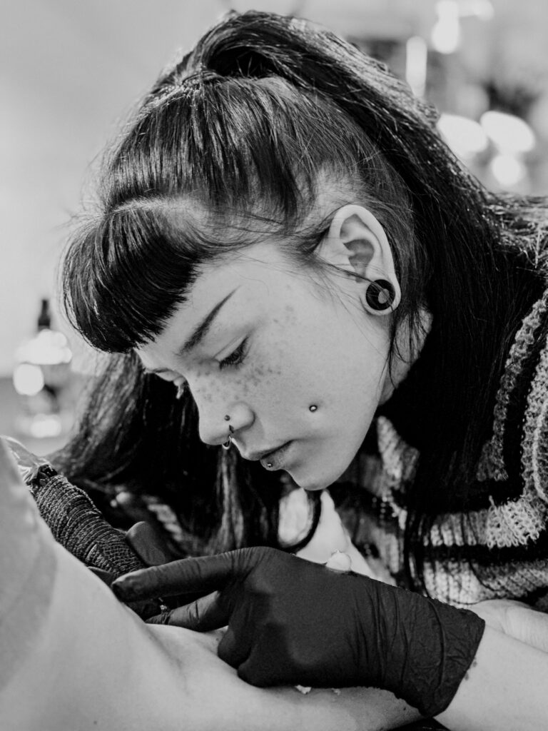 A black and white close-up of a tattoo artist with dark hair in a high ponytail and multiple facial piercings. Wearing black gloves, the artist is deeply focused while working on a tattoo on a client's arm.