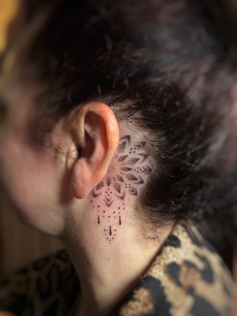 A delicate and precise dotwork tattoo placed behind a person's ear, created by Celia from Poppy Lia. The design is a small, ornate mandala featuring fine-point details and dangling ornamental accents.