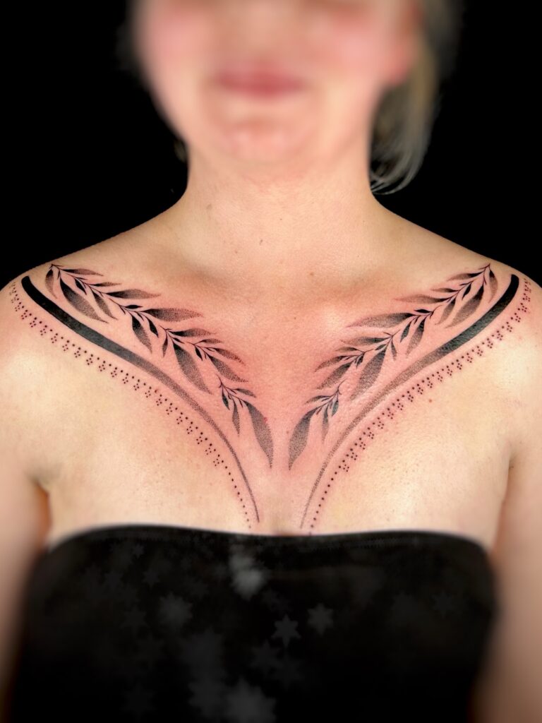 A graceful and symmetrical botanical tattoo on a client's upper chest and collarbone area by Celia of Poppy Lia. The design features elegant, leafy vines extending outward, rendered with fine lines and soft shading.