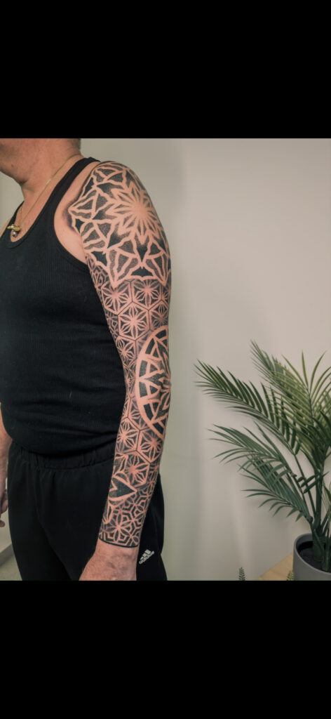 A comprehensive black-ink geometric sleeve tattoo on an arm by Carlo of Carlo Tattoo. The design features various patterns, including large mandalas on the shoulder and elbow connected by intricate repeating hexagonal and star shapes.