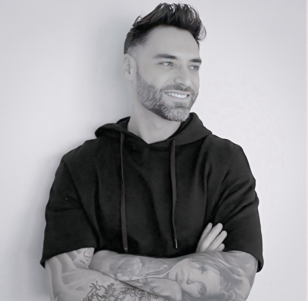 A black and white portrait of tattoo artist Carlo from Carlo Tattoo. He has a groomed beard and styled hair, wearing a black hoodie and posing with his tattooed arms crossed while looking to the side with a smile.