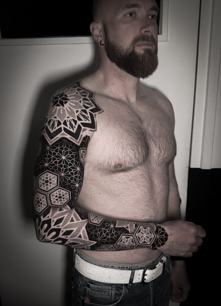A striking, high-contrast black-and-grey sleeve tattoo by Andy of Deux Tattoo. The design features intricate mandalas, floral patterns, and interlocking geometric hexagons covering the entire arm and hand.