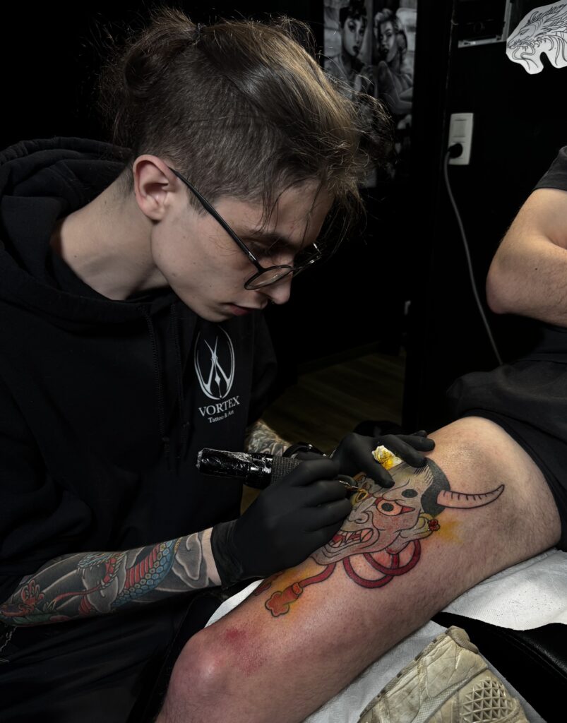 A close-up of tattoo artist Yento from Yentonic Tattoo at work. He is wearing glasses and a black "Vortex" hoodie, deeply focused on tattooing a colorful Japanese Hannya mask onto a client's thigh.