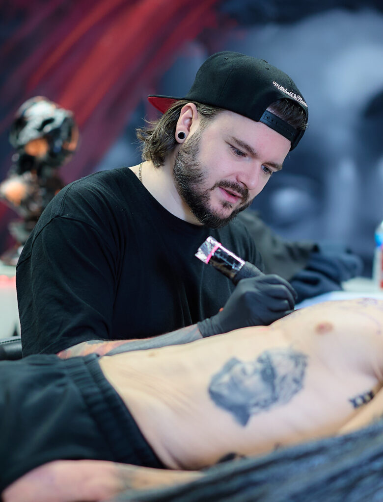 A photograph of tattoo artist Kenny from Hannon Tattoo deeply focused on his work. He has a beard and is wearing a black t-shirt and a black baseball cap backward while using a tattoo machine on a client's torso.