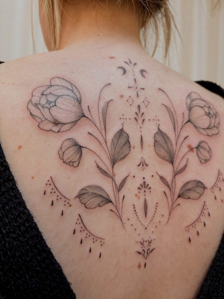 A close-up shot of a fine-line back tattoo by Tacha from Studio Manoja, highlighting the precise linework, soft shading on the petals, and the perfect symmetry of the ornamental design.