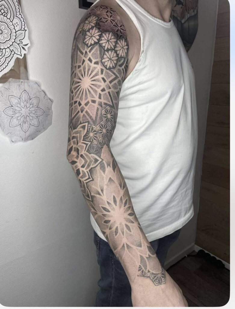 A complex black-and-grey geometric sleeve tattoo on an arm, expertly rendered by Emmi of Pink Flamingo. The piece features various interlocking star-like and floral geometric patterns using fine dotwork for depth.