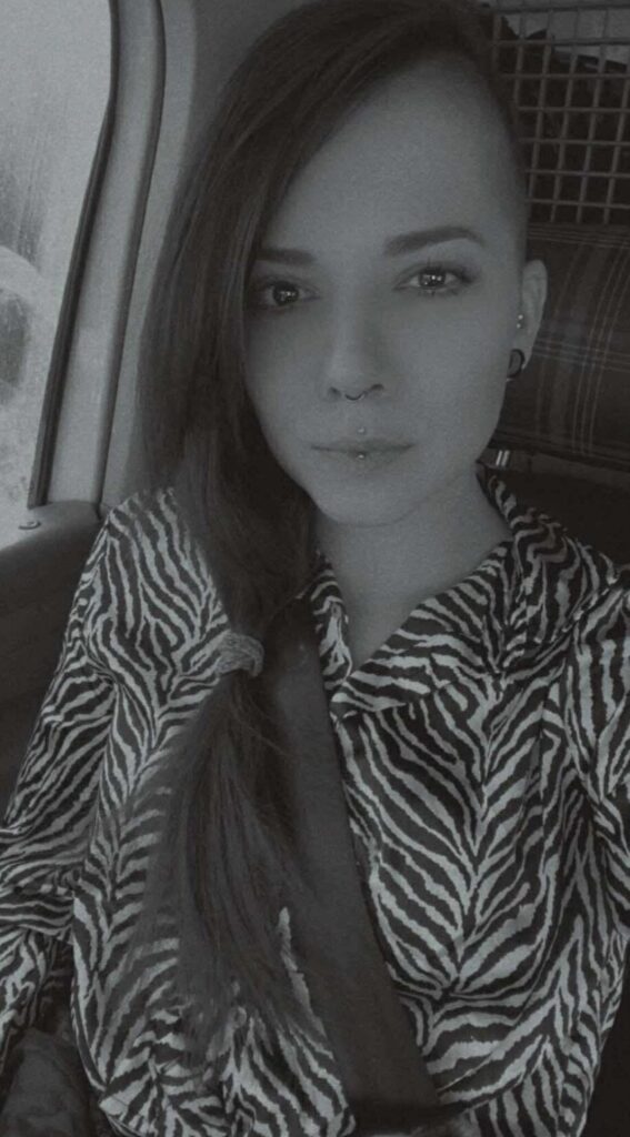 A black and white portrait of tattoo artist Emmi from Pink Flamingo. She has long hair swept to one side with a shaved side, a septum piercing, and a vertical labret piercing.