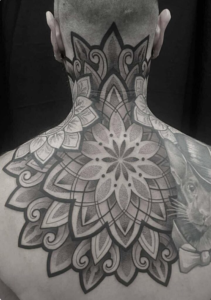A large, symmetrical black-and-grey mandala tattoo on a client's upper back and neck, created by Emmi of Pink Flamingo. The intricate design features layers of ornamental patterns and delicate dotwork shading.
