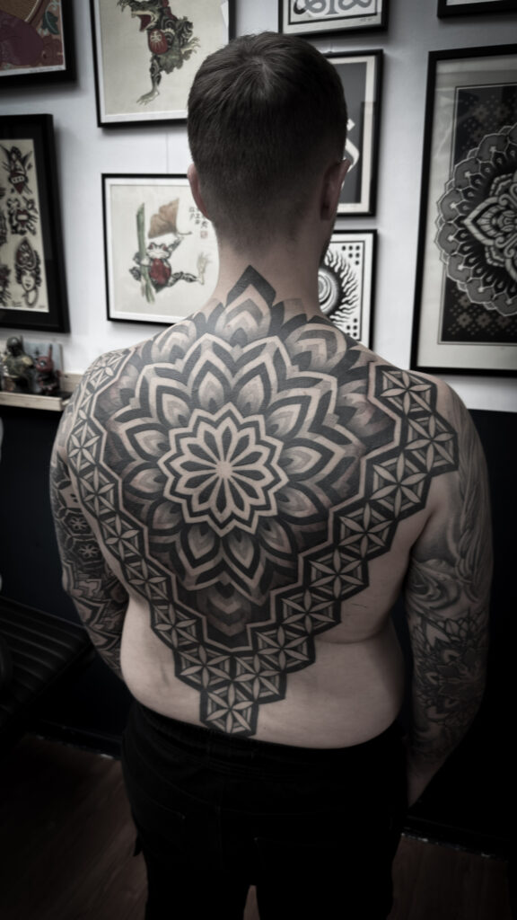 A massive, symmetrical black-ink mandala tattoo covering a man's entire back, created by Andy of Deux Tattoo. The design is centered on the spine and expands outward into sharp, ornamental patterns and geometric "Flower of Life" details.