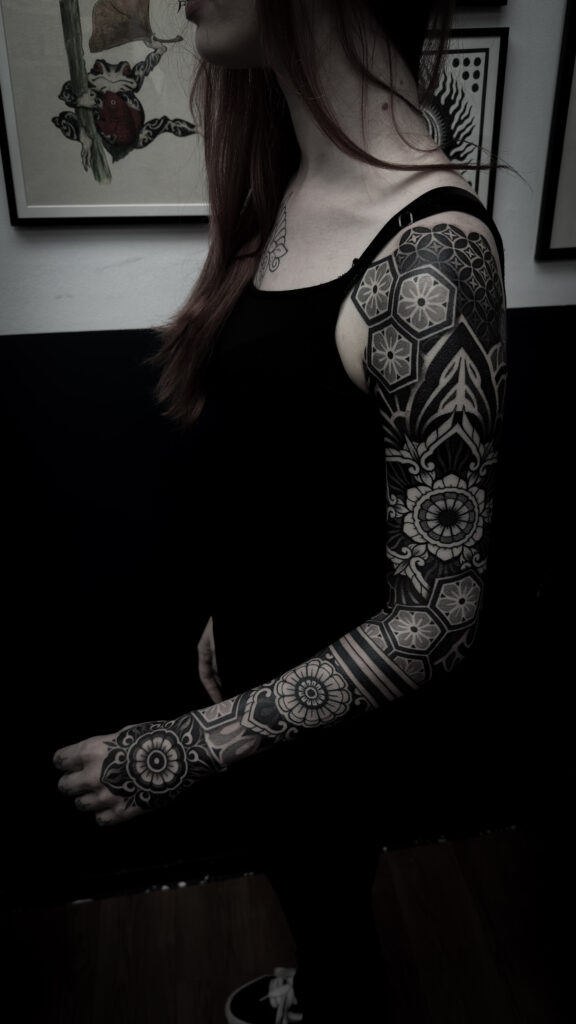 A bold, full-sleeve geometric tattoo on a man's arm by Andy from Deux Tattoo. It features large, dark star shapes and hexagonal "honeycomb" patterns filled with complex, detailed dotwork.