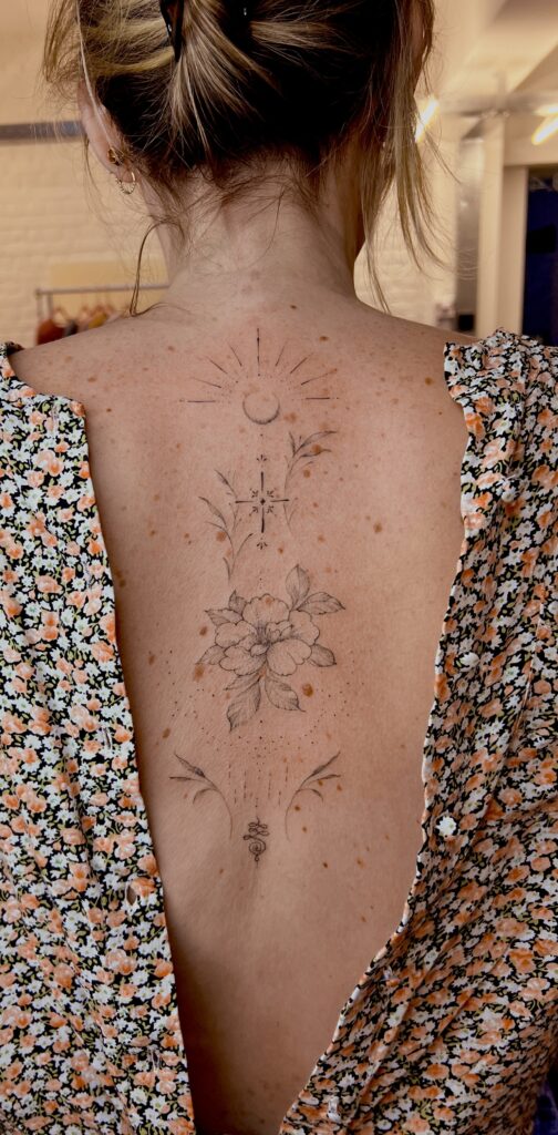 A vertical, fine-line ornamental tattoo placed along the spine, created by Tacha of Studio Manoja. The design incorporates a central flower with celestial elements like a crescent moon, a sunburst, and elegant flourishes.