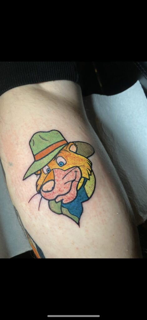 A vibrant, illustrative color tattoo by Delfien of Trick or Tat depicting a classic cartoon character wearing a green hat with an orange band and a blue bandana.