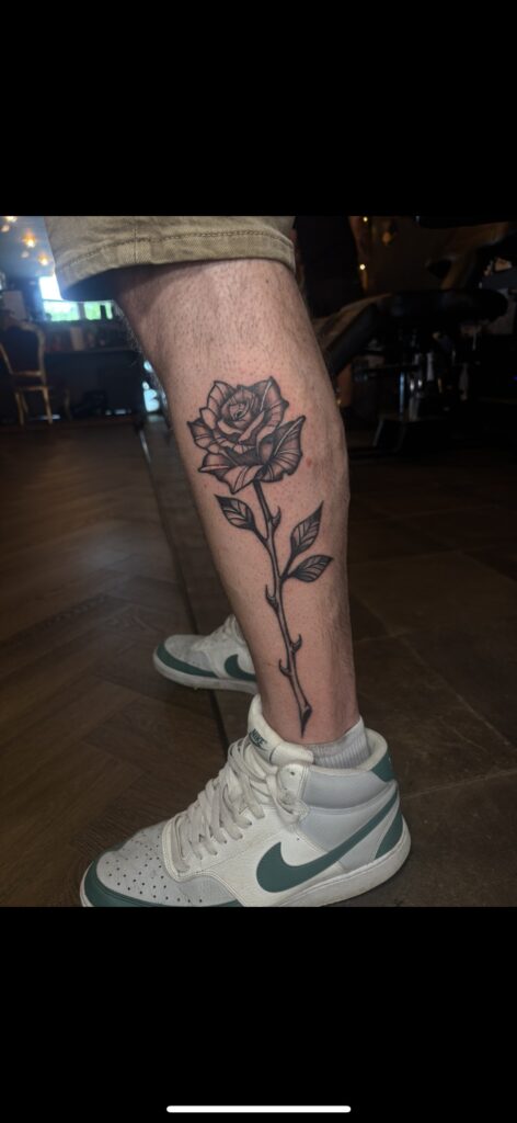 A detailed black and grey tattoo of a single rose with a long, thorny stem on a calf, showcasing the clean linework and shading of Delfien from Trick or Tat.