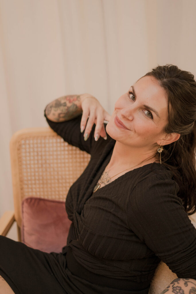 A portrait of tattoo artist Tacha from Studio Manoja sitting comfortably in a wicker chair and smiling. She is wearing a black wrap-style dress, and her arms are covered in her own colorful illustrative and floral tattoos.