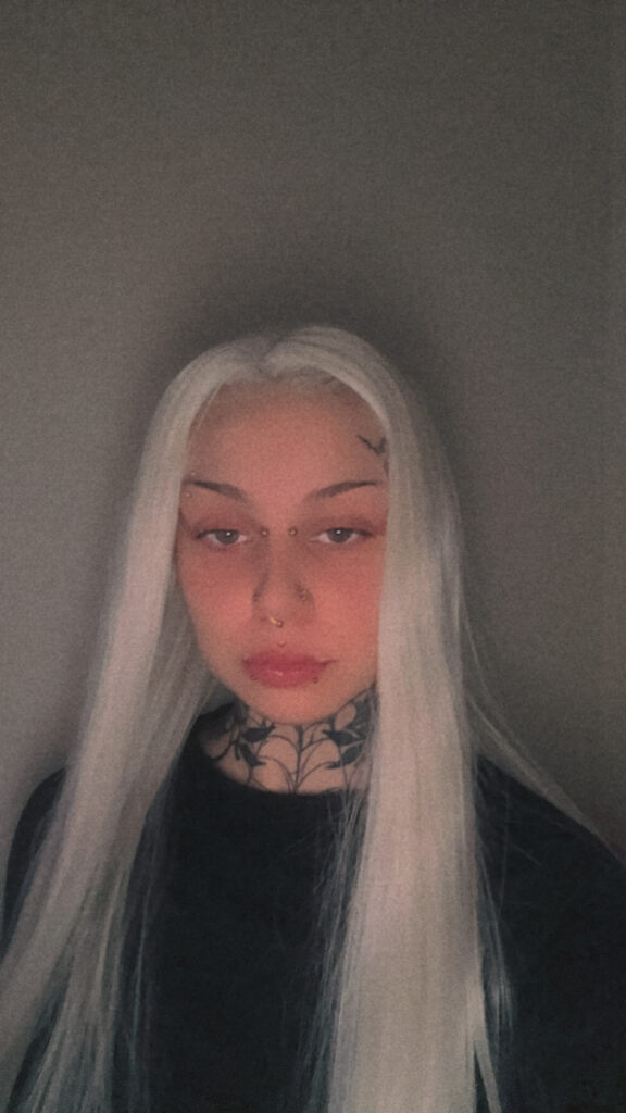 A portrait of tattoo artist Delfien from Trick or Tat, featuring her long white hair and multiple facial piercings. A bold blackwork tattoo of a spiderweb and floral patterns covers her neck.