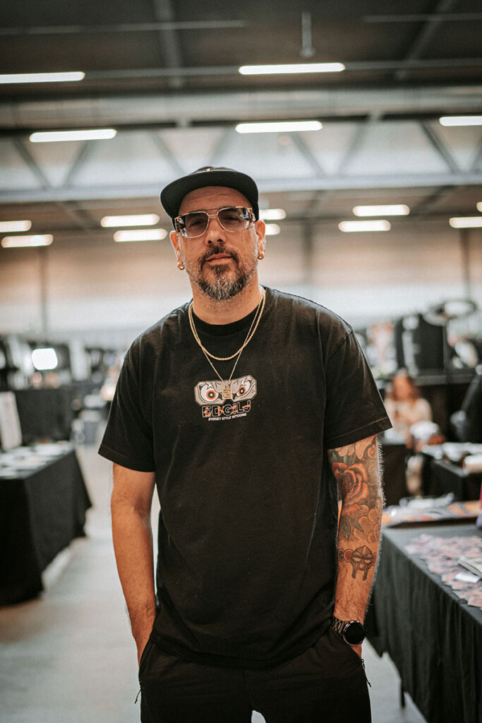 A portrait of tattoo artist Quincy from Kwillsink at a convention. He is wearing a black t-shirt with a graphic logo, gold chains, a black baseball cap, and tinted glasses. He has a groomed beard and tattoos are visible on his left arm.