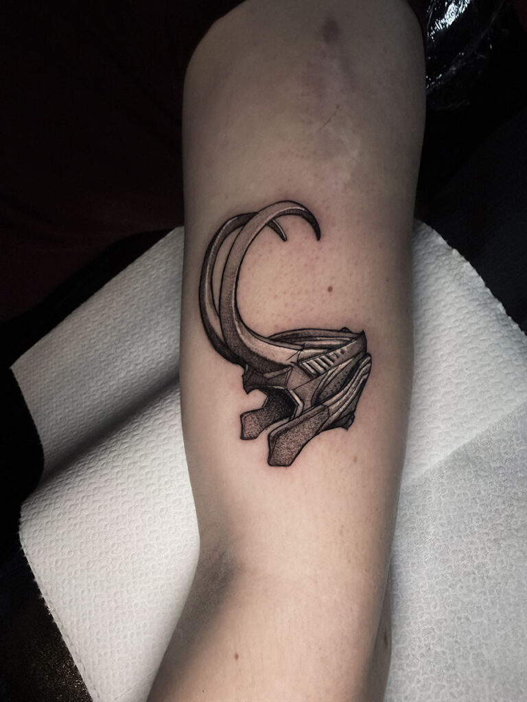 A detailed black and grey tattoo on a limb by Quincy of Kwillsink, depicting the iconic horned helmet worn by the character Loki. The design features clean linework and smooth dotwork shading to give the helmet a metallic appearance.
