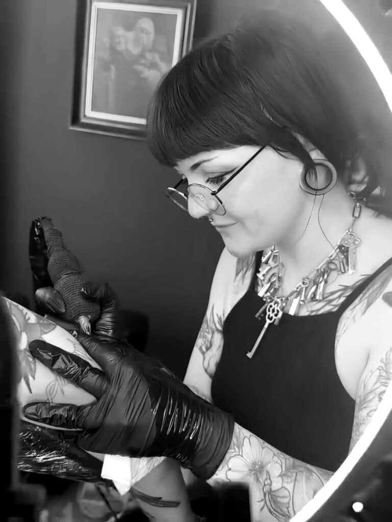 A black and white photo of tattoo artist Najate from Empowering Life at work. She is wearing glasses and a unique necklace of various keys, focused intently while tattooing a client's arm.