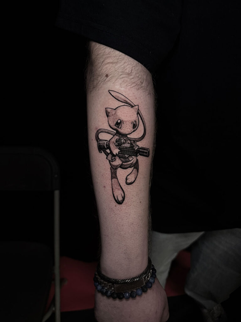 A unique black and grey forearm tattoo by Quincy of Kwillsink featuring the Pokémon Mew. The character is rendered with soft shading while holding a large, highly detailed tactical rifle, creating a contrast between "cute" and "gritty."