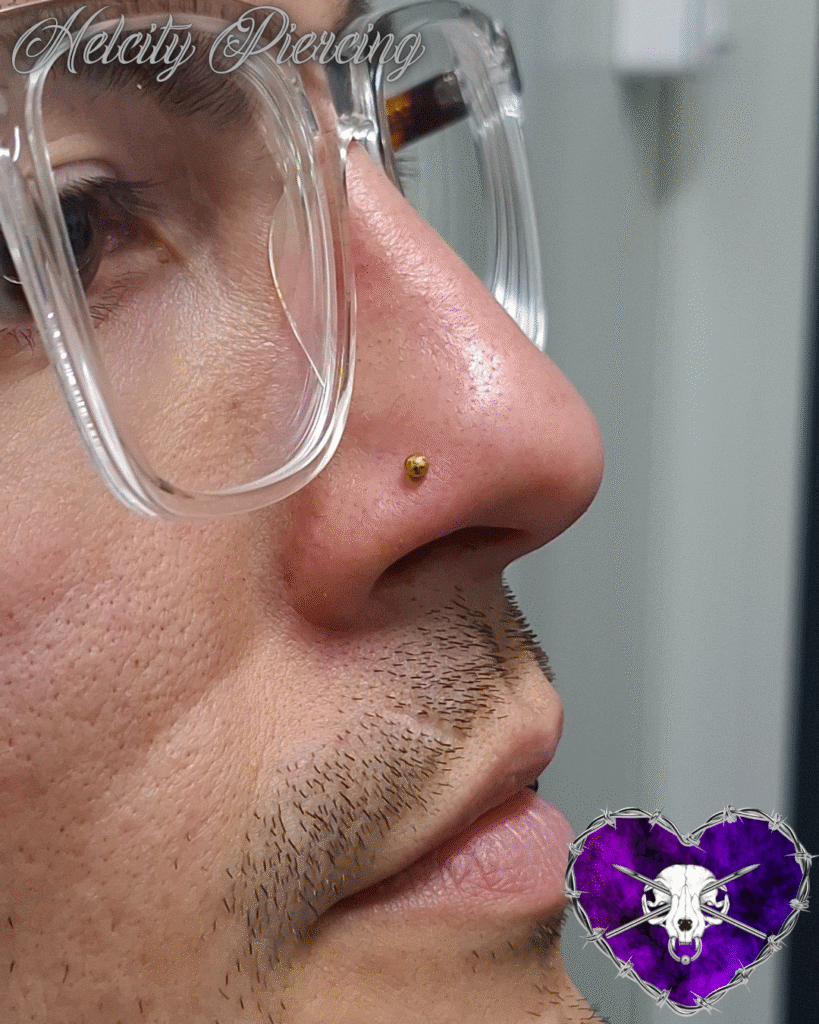 A profile close-up of a person wearing clear-framed glasses, highlighting a clean and classic gold ball stud nostril piercing expertly placed by Milanda of Helcity Piercing.